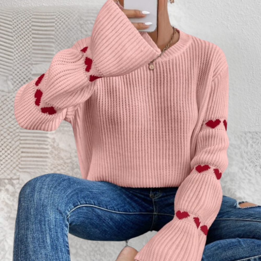 Vrouw draagt roze trui met rode hartjes op mouwen, gecombineerd met blauwe jeans. Modieuze dameskleding, casual outfit, herfststijl.