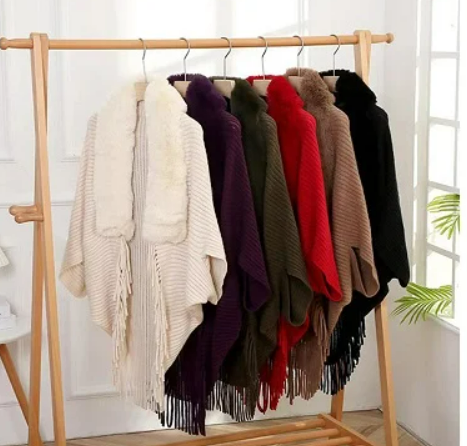 Poncho's met franjes in diverse kleuren, zoals wit, paars, groen, rood, bruin en zwart, hangen op een houten kledingrek in een lichte kamer.