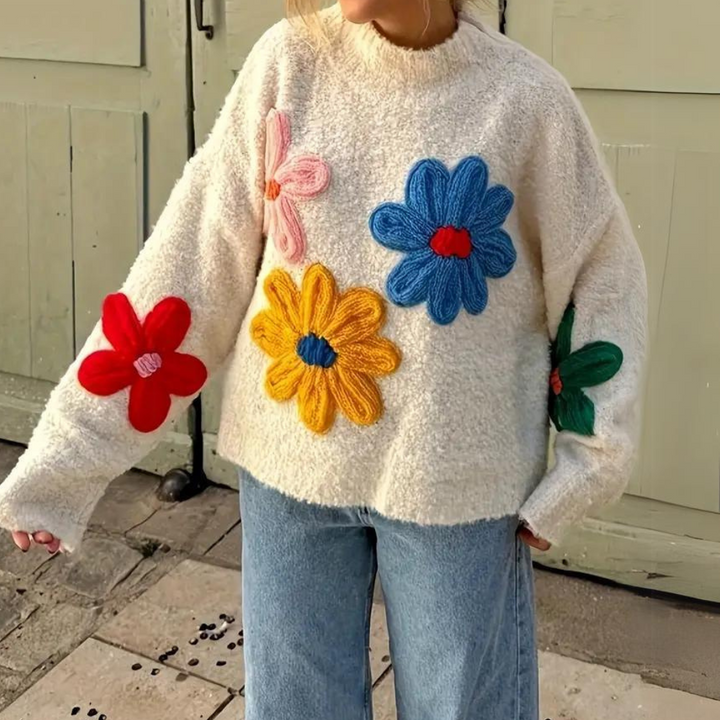 Vrouw draagt een crèmekleurige trui met kleurrijke geborduurde bloemen, gecombineerd met blauwe jeans. Trendy herfstmode, casual en stijlvol.