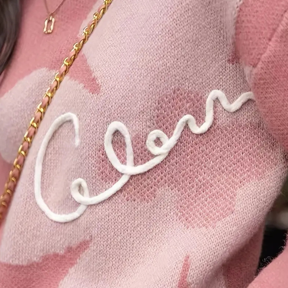 Roze trui met witte geborduurde tekst "Celine", gouden ketting, modieuze dameskleding, trendy knitwear, herfstmode, close-up detail.