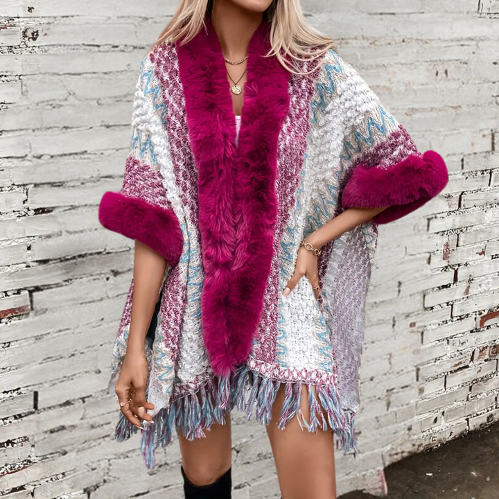 Vrouw draagt kleurrijke poncho met roze bontkraag en franjes, tegen witte bakstenen muur. Modieuze winterkleding, trendy damesmode.