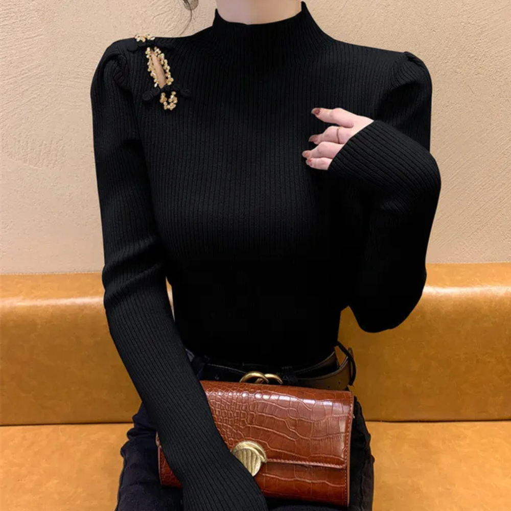 Zwarte geribbelde coltrui met lange mouwen, gouden schouderdetails, gecombineerd met bruine krokodillenleren clutch op gele bank.