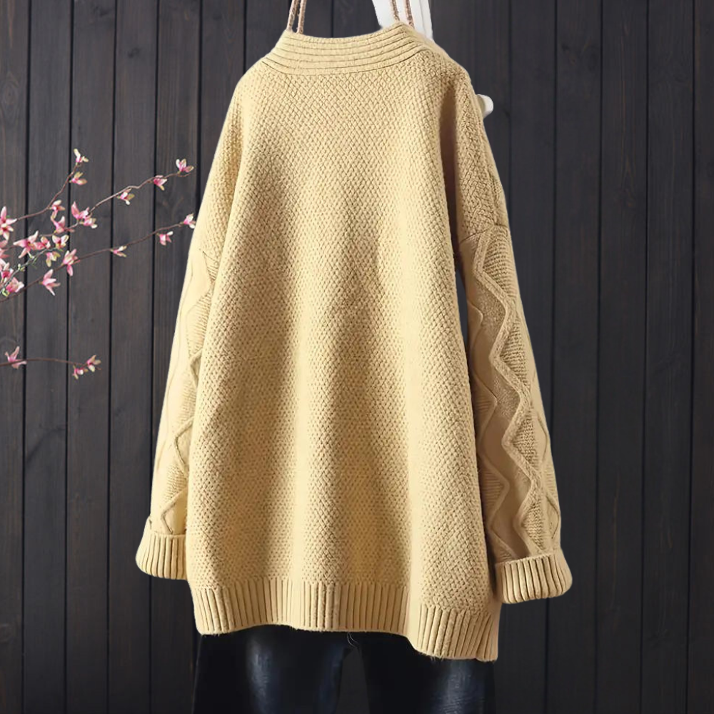 Beige oversized gebreide trui met textuurpatroon, lange mouwen en ronde hals, hangend voor een donkere houten achtergrond. Perfect voor herfstmode.
