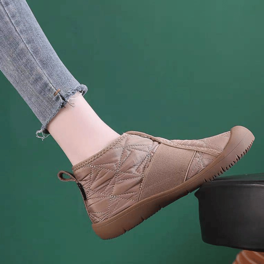 Beige gewatteerde pantoffel met rubberen zool, gedragen door persoon in blauwe jeans, tegen groene achtergrond. Comfortabele indoor schoenen.