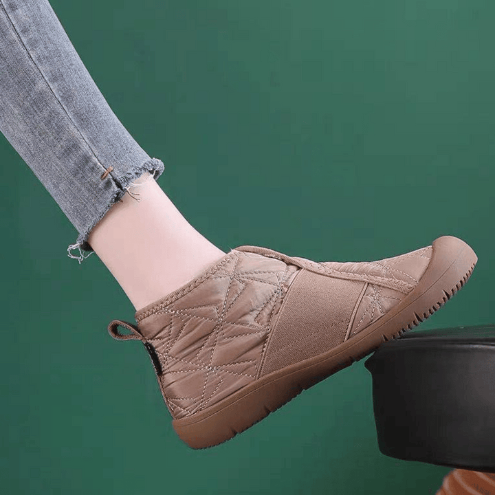 Beige gewatteerde pantoffel met rubberen zool, gedragen door persoon in blauwe jeans, tegen groene achtergrond. Comfortabele indoor schoenen.