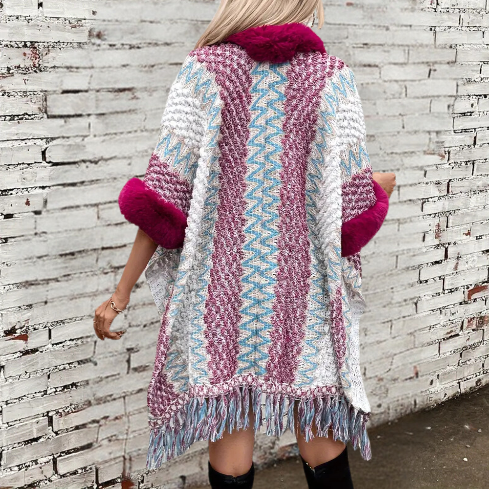 Vrouw in kleurrijke gebreide poncho met franjes en roze accenten, tegen een witte bakstenen muur. Modieuze herfstkleding, boho stijl.