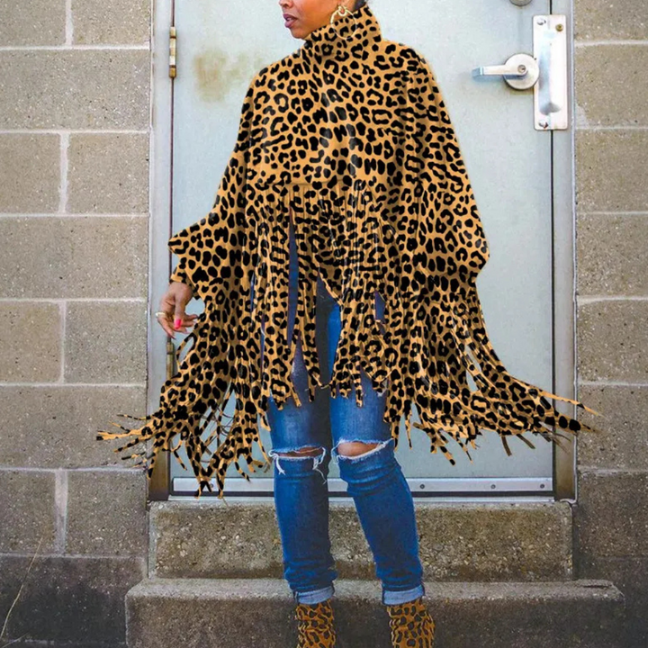 Vrouw draagt een luipaardprint poncho met franjes, gecombineerd met gescheurde jeans en laarzen, staand voor een betonnen muur.