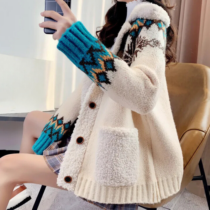 Vrouw in beige gebreid vest met blauwe en bruine patronen, zittend op stoel, houdt smartphone vast. Wintermode, comfortabele kleding, trendy design.