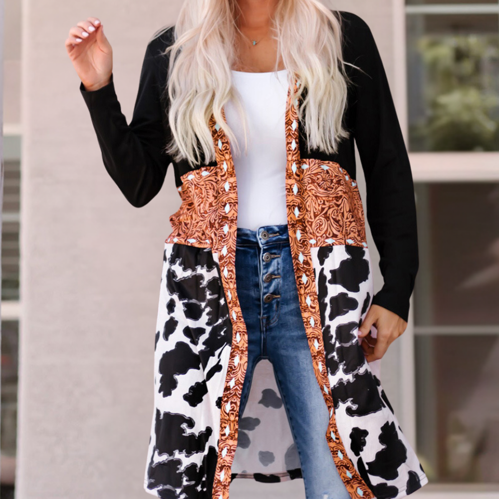 Vrouw in modieuze lange jas met koeienprint en oranje patroon, gecombineerd met blauwe jeans en wit shirt, buiten voor een gebouw.
