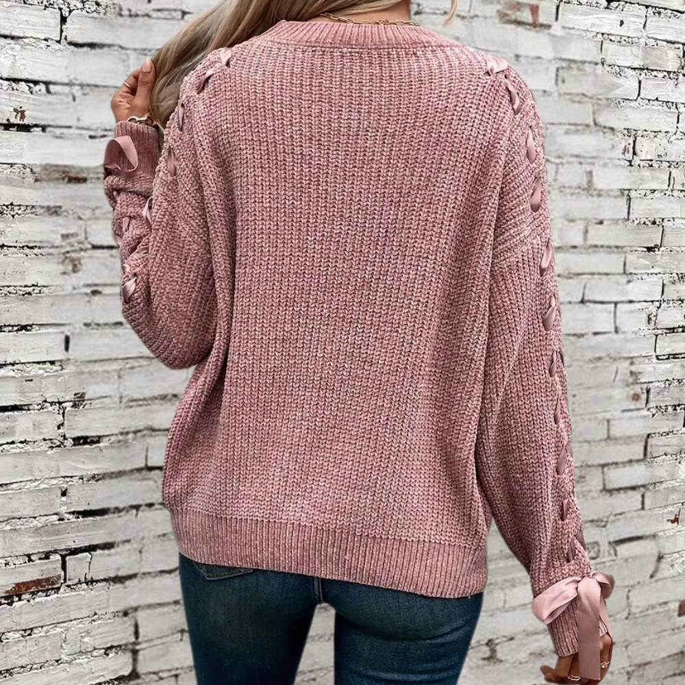 Vrouw in roze gebreide trui met vlechtpatroon en strikdetails, staand voor witte bakstenen muur. Modieuze dameskleding, herfststijl.
