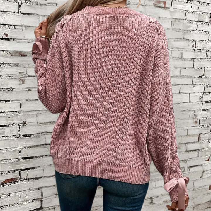 Vrouw in roze gebreide trui met vlechtpatroon en strikdetails, staand voor witte bakstenen muur. Modieuze dameskleding, herfststijl.