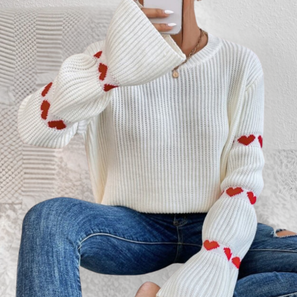 Vrouw in witte trui met rode hartjes op mouwen, zittend in spijkerbroek, houdt smartphone vast. Modieuze dameskleding, casual winteroutfit.