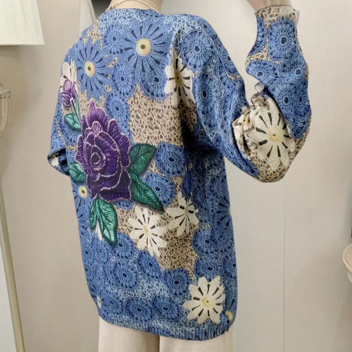 Blauwe gebreide trui met kleurrijke bloemenprint, waaronder een grote paarse roos en witte madeliefjes. Modieuze dameskleding, herfststijl.