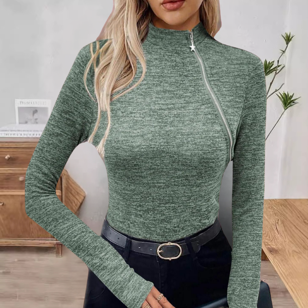 Vrouw in groene coltrui met ritssluiting, lange mouwen, zwarte broek en riem, staand voor houten kast en plant. Modieuze dameskleding.
