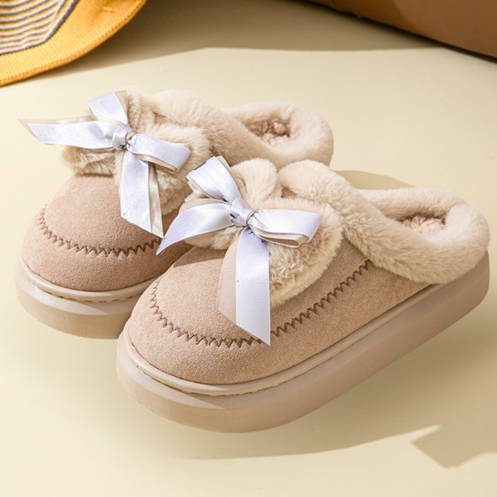 Beige damespantoffels met pluche voering en witte strik, comfortabele winterse huisschoenen, antislip zool, perfect voor binnengebruik.