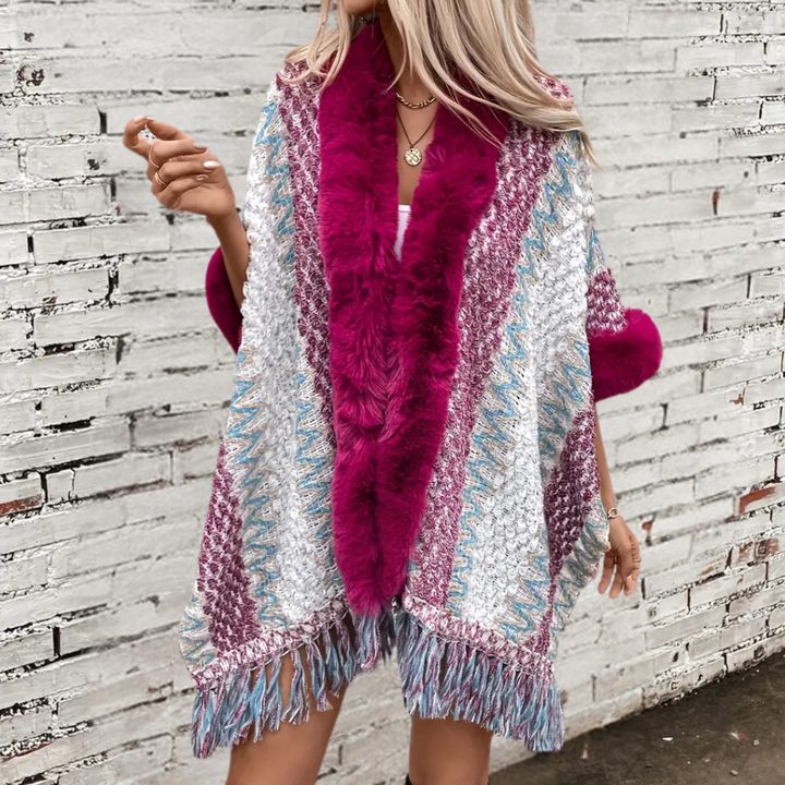 Vrouw draagt kleurrijke poncho met roze bontkraag en franjes, tegen witte bakstenen muur. Modieuze dameskleding, herfststijl, boho-chic.