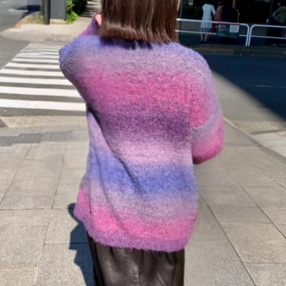 Vrouw in kleurrijke, gebreide trui met roze en paarse strepen, loopt op trottoir bij zebrapad. Modieuze herfstkleding, stedelijke omgeving.