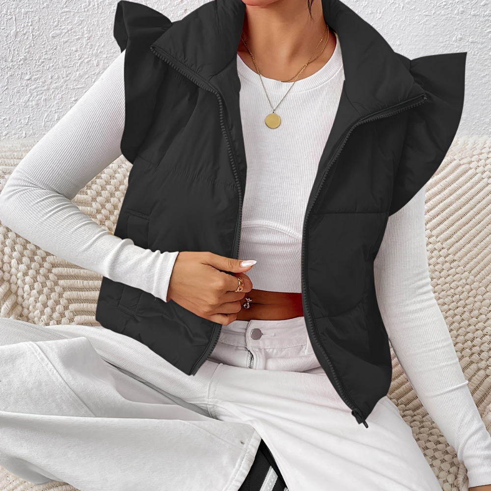 Vrouw draagt zwarte gewatteerde bodywarmer met rits en ruches, gecombineerd met witte trui en broek. Modieuze dameskleding, herfst/winter stijl.