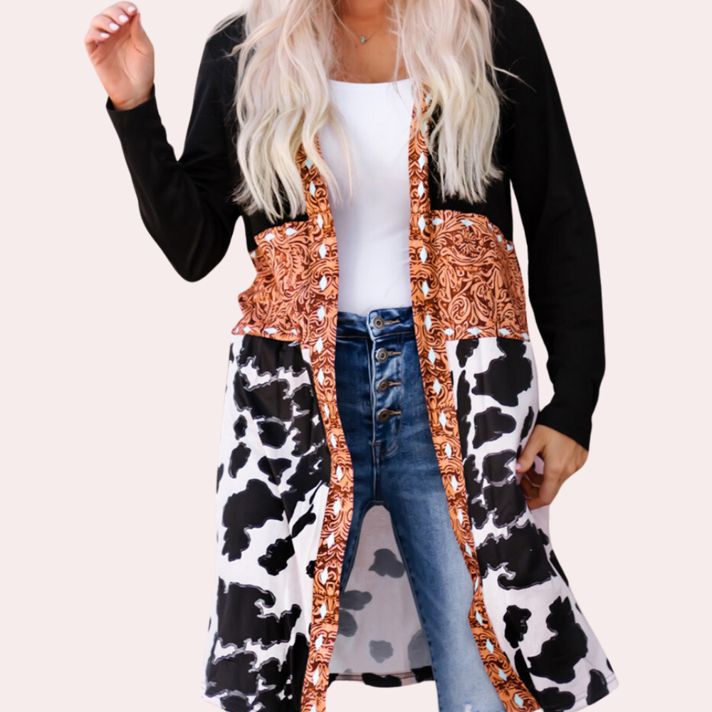 Vrouw draagt lange cardigan met koeienprint en oranje paisley-patroon, gecombineerd met een witte top en blauwe jeans. Modieuze dameskleding.