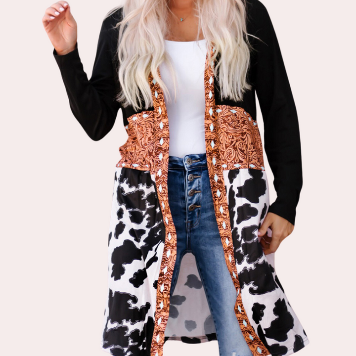 Vrouw draagt lange cardigan met koeienprint en oranje paisley-patroon, gecombineerd met een witte top en blauwe jeans. Modieuze dameskleding.