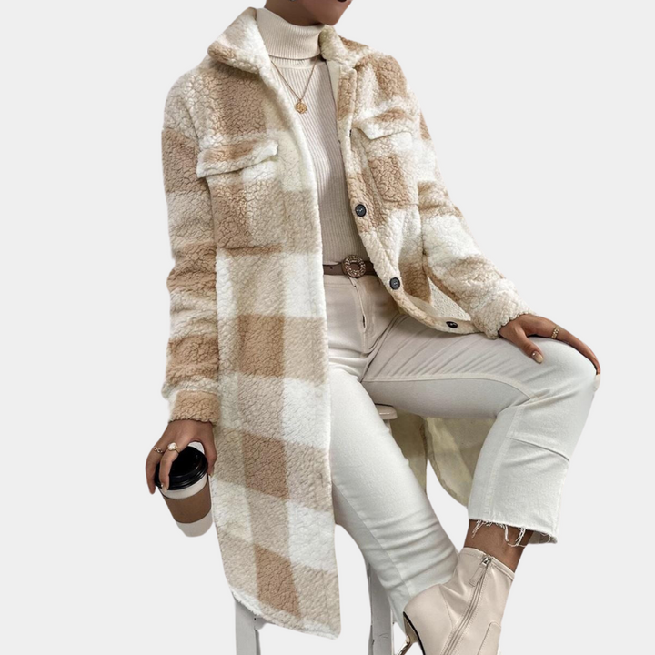 Vrouw draagt lange geruite teddyjas in beige en wit, gecombineerd met witte coltrui en broek. Modieuze herfst- of winteroutfit met enkellaarsjes.