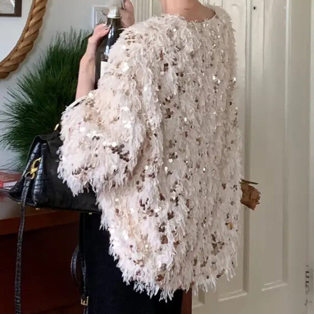 Dame in glinsterende paillettenjas met zwarte handtas, staand voor een witte deur. Modieuze feestkleding, trendy damesmode, luxe uitstraling.