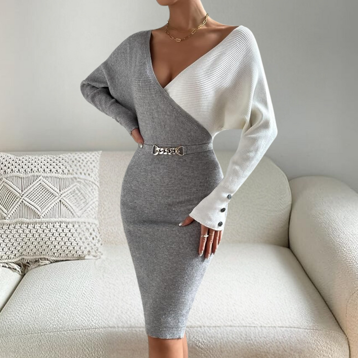 Grijze en witte gebreide jurk met V-hals en riem, gedragen door een model op een crèmekleurige bank. Elegante damesmode, herfst-winter stijl.