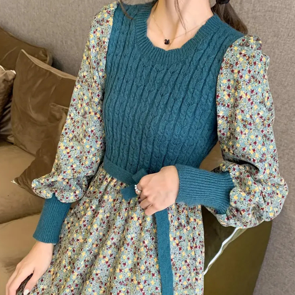 Vrouw in elegante jurk met gebreide blauwe top en bloemenprint rok, lange pofmouwen, trendy mode, comfortabele bank op achtergrond.
