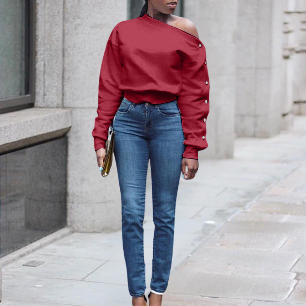 Vrouw in rode off-shoulder trui en blauwe skinny jeans, staand op trottoir. Stijlvolle mode, casual outfit, stedelijke achtergrond.