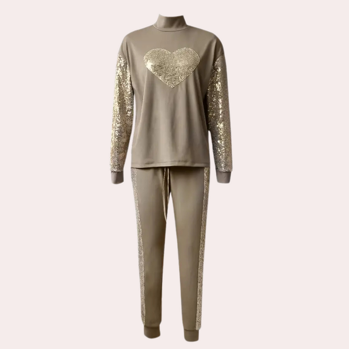 Beige dames trainingspak met gouden pailletten, hartmotief op de borst en glinsterende mouwen. Modieuze loungewear, comfortabele pasvorm.