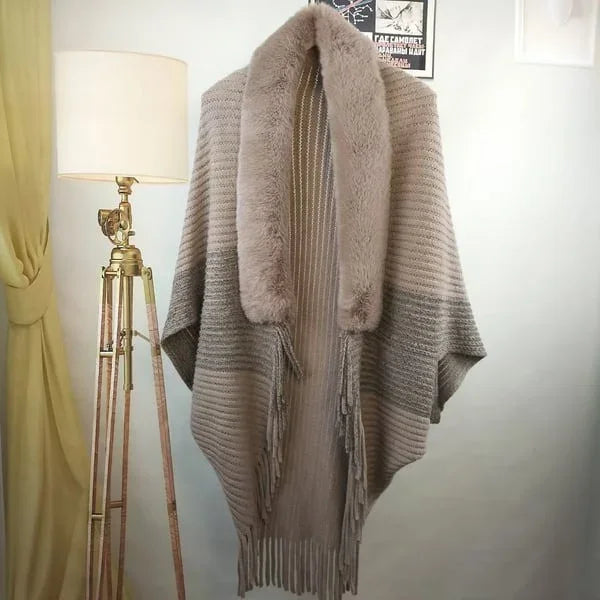 Khaki poncho met bontkraag, franjes en geribbelde textuur, hangend naast een vintage vloerlamp. Stijlvolle damesmode, herfst/winter outfit.