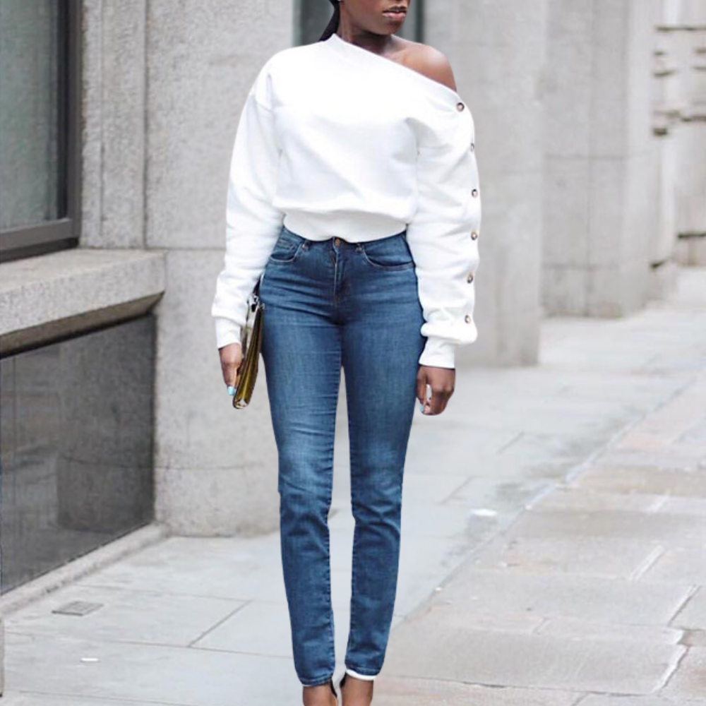 Vrouw in witte off-shoulder trui en blauwe skinny jeans, staand op trottoir. Modieuze casual outfit, stedelijke achtergrond.