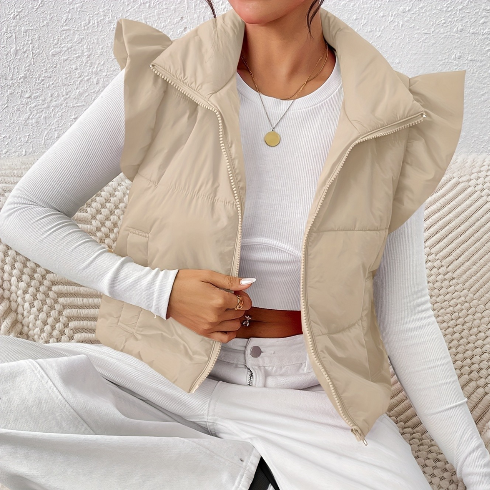 Vrouw draagt beige gewatteerd vest met ruches, witte lange mouwen top en grijze broek. Modieuze winterkleding, casual stijl, comfortabele outfit.