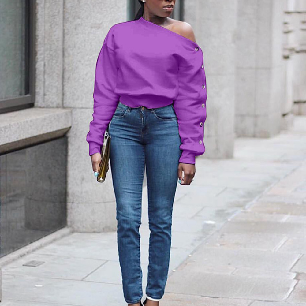 Vrouw in paarse off-shoulder trui en blauwe jeans, staand op trottoir. Modieuze casual outfit, stedelijke achtergrond.