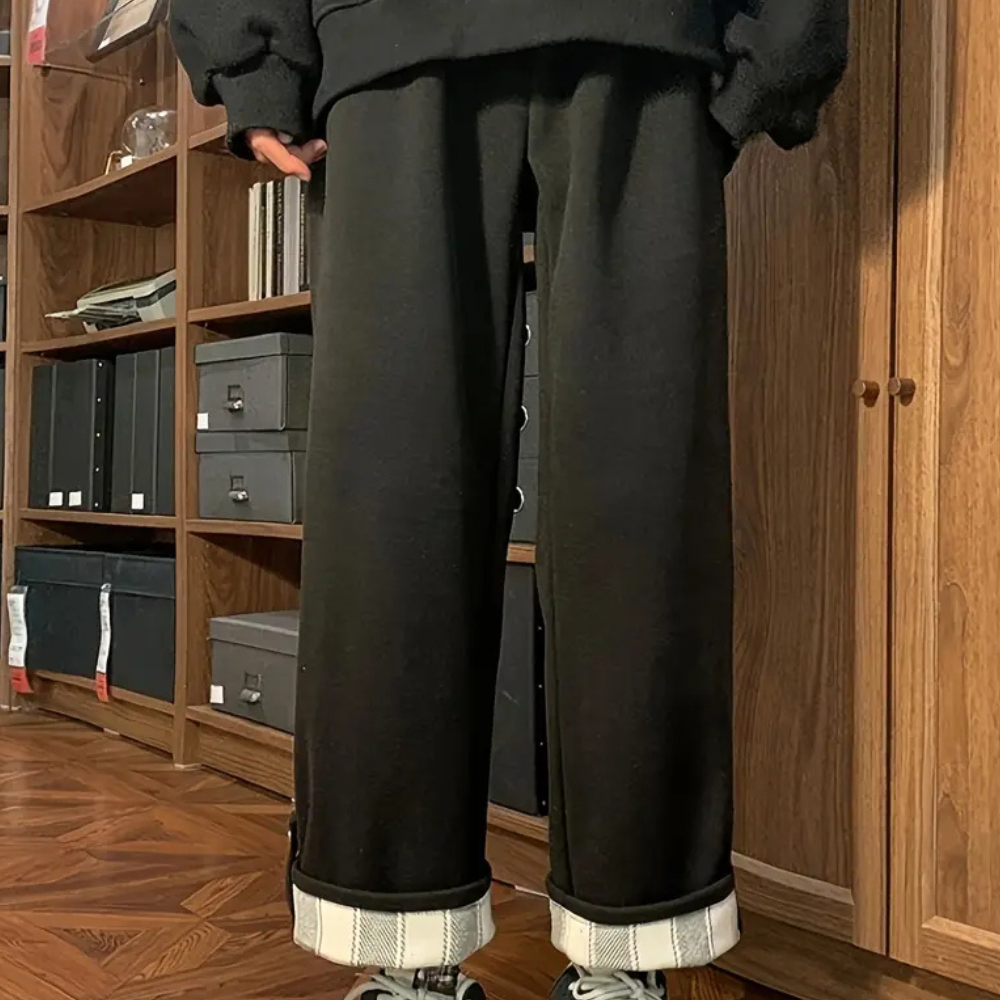 Zwarte broek met opgerolde pijpen, gedragen door persoon in moderne kamer met houten vloer en boekenplanken. Comfortabele, casual kledingstijl.