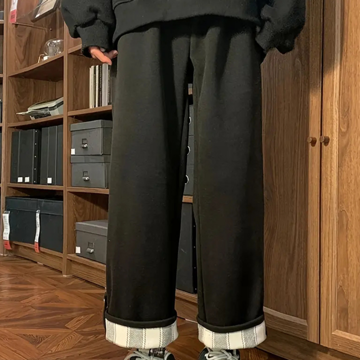 Zwarte broek met opgerolde pijpen, gedragen door persoon in moderne kamer met houten vloer en boekenplanken. Comfortabele, casual kledingstijl.