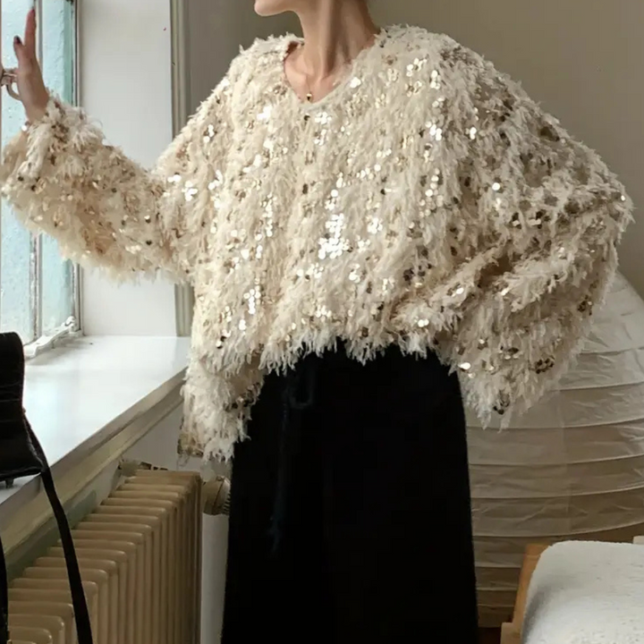 Vrouw draagt een beige, pluizige trui met pailletten, staand bij een raam. Modieuze dameskleding, herfst-winter stijl, trendy oversized look.