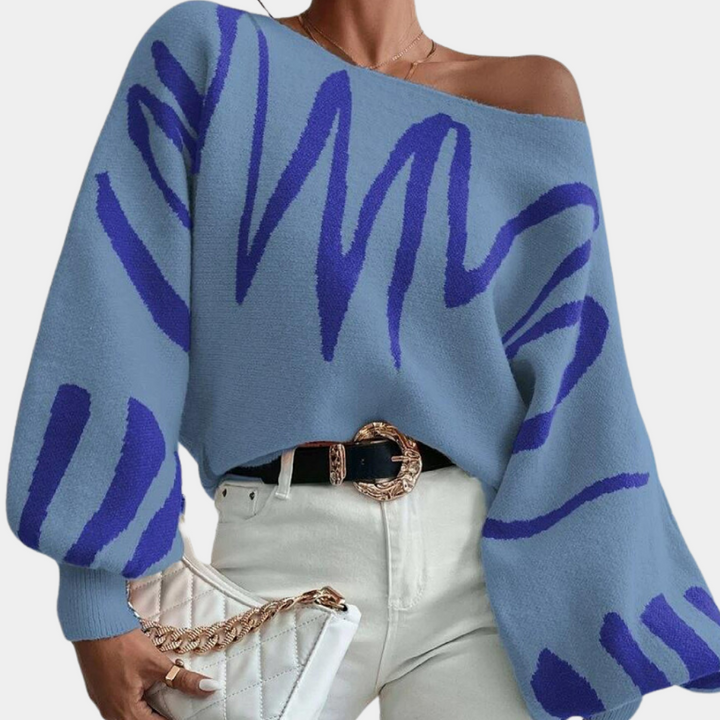 Vrouw draagt blauwe trui met abstract patroon, off-shoulder ontwerp. Modieuze dameskleding, casual stijl, gecombineerd met witte broek en gewatteerde tas.