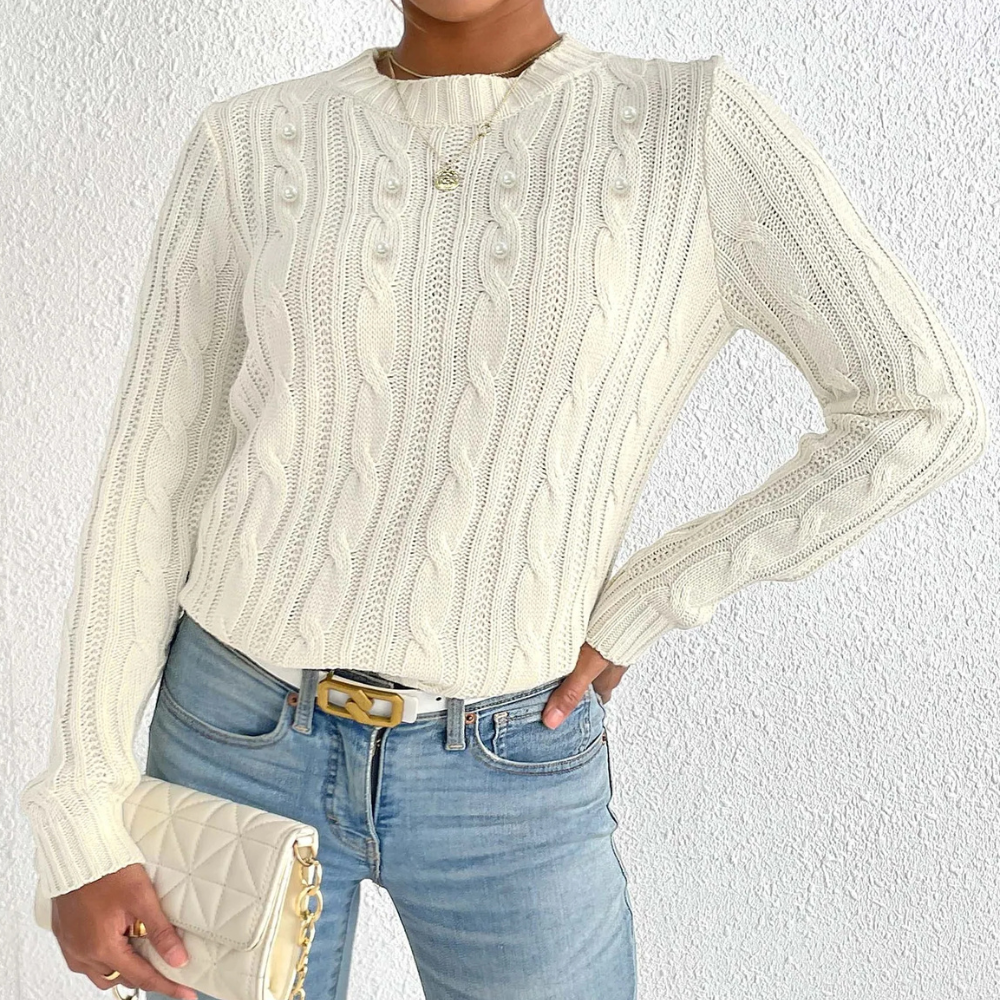 Vrouw in crèmekleurige kabeltrui met gouden ketting, blauwe jeans en gewatteerde clutch. Modieuze dameskleding, casual stijl.
