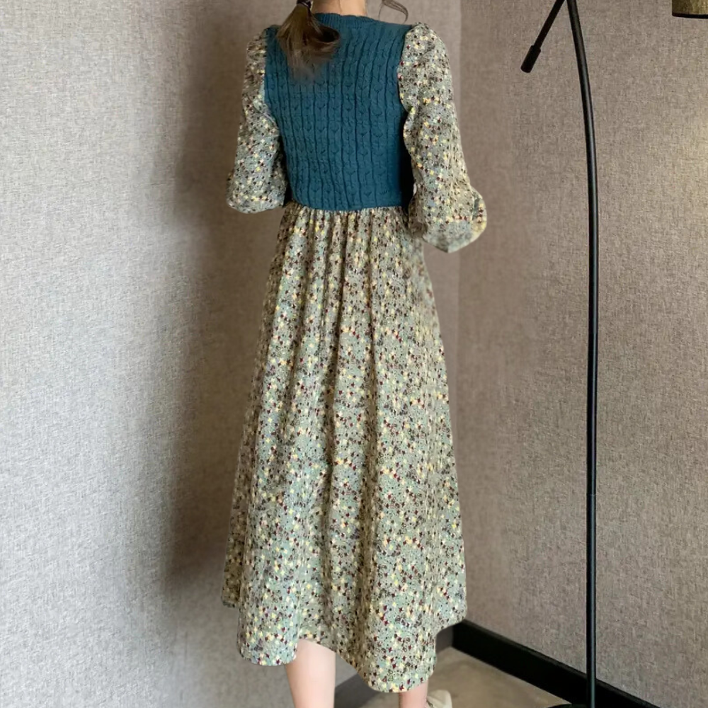 Vrouw in bloemenjurk met gebreid blauw bovenstuk, lange mouwen, staande in moderne kamer. Vintage mode, dameskleding, herfststijl.
