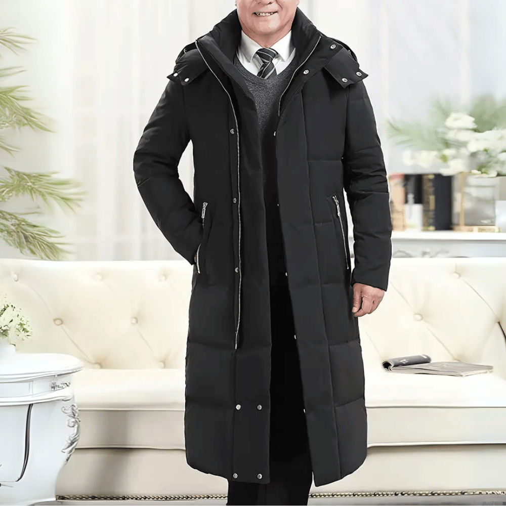 Zwarte lange winterjas voor heren met capuchon, ritssluiting en zakken, gedragen in een stijlvolle woonkamer. Perfect voor koude seizoenen.