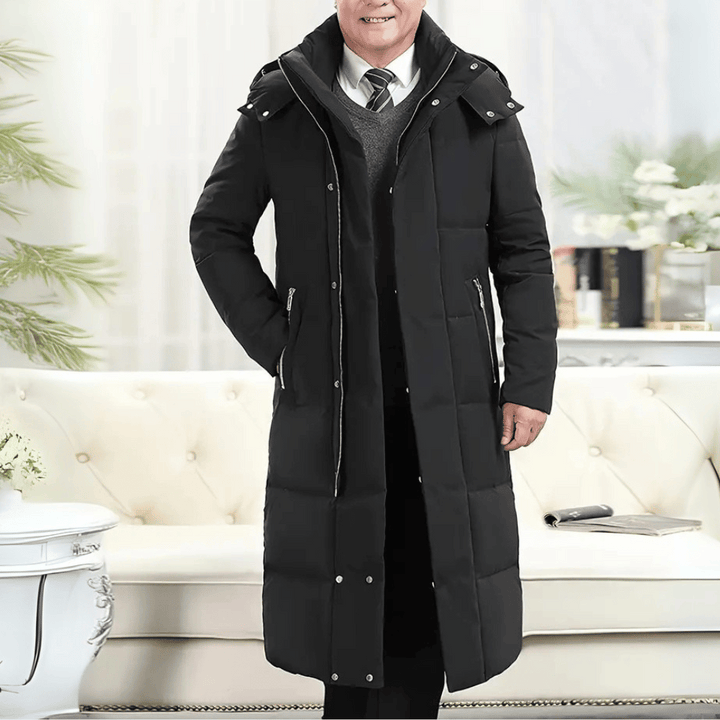 Zwarte lange winterjas voor heren met capuchon, ritssluiting en zakken, gedragen in een stijlvolle woonkamer. Perfect voor koude seizoenen.
