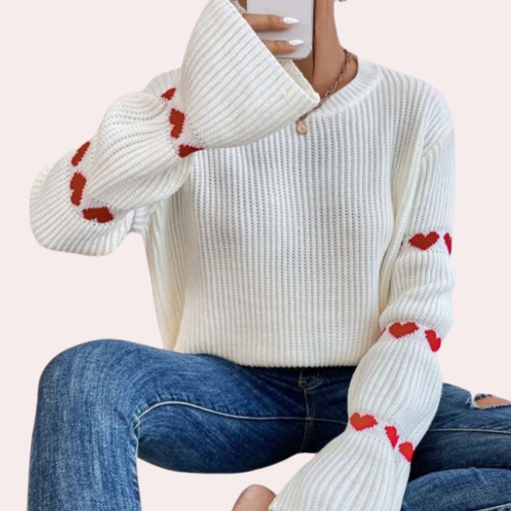 Vrouw in witte trui met rode hartjes, zittend in spijkerbroek, drinkt koffie. Modieuze dameskleding, casual winteroutfit, trendy stijl.