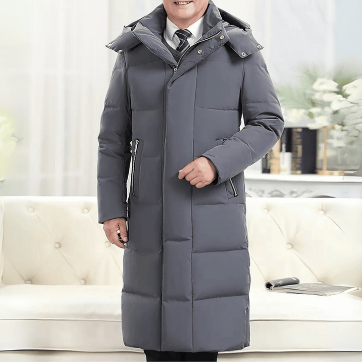 Grijze lange winterjas voor heren, gewatteerd met capuchon, gedragen door een man in een moderne woonkamer. Stijlvolle en warme herenmode.