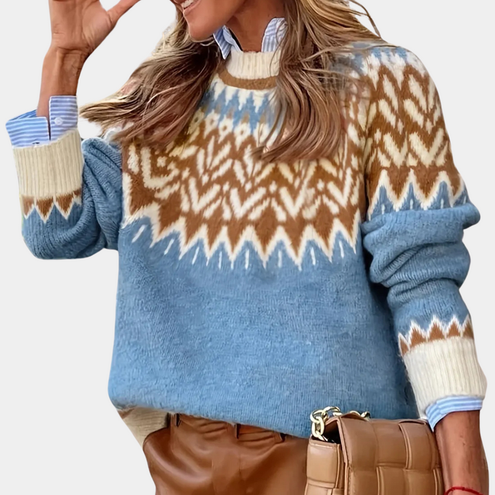 Vrouw in blauwe Noorse trui met bruin-wit patroon, draagt leren broek en gevlochten tas. Modieuze winterkleding, Scandinavisch design.