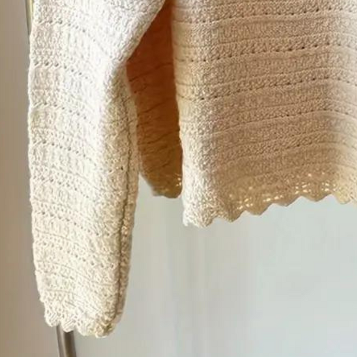 Crèmekleurige gebreide trui met lange mouwen, gedetailleerd patroon, hangend aan een kledingrek. Perfect voor herfstmode en casual outfits.
