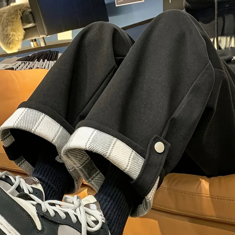Zwarte wijde broek met geruite omslag, gedragen met zwarte sokken en sneakers, op een bruine leren bank. Modieuze casual outfit.