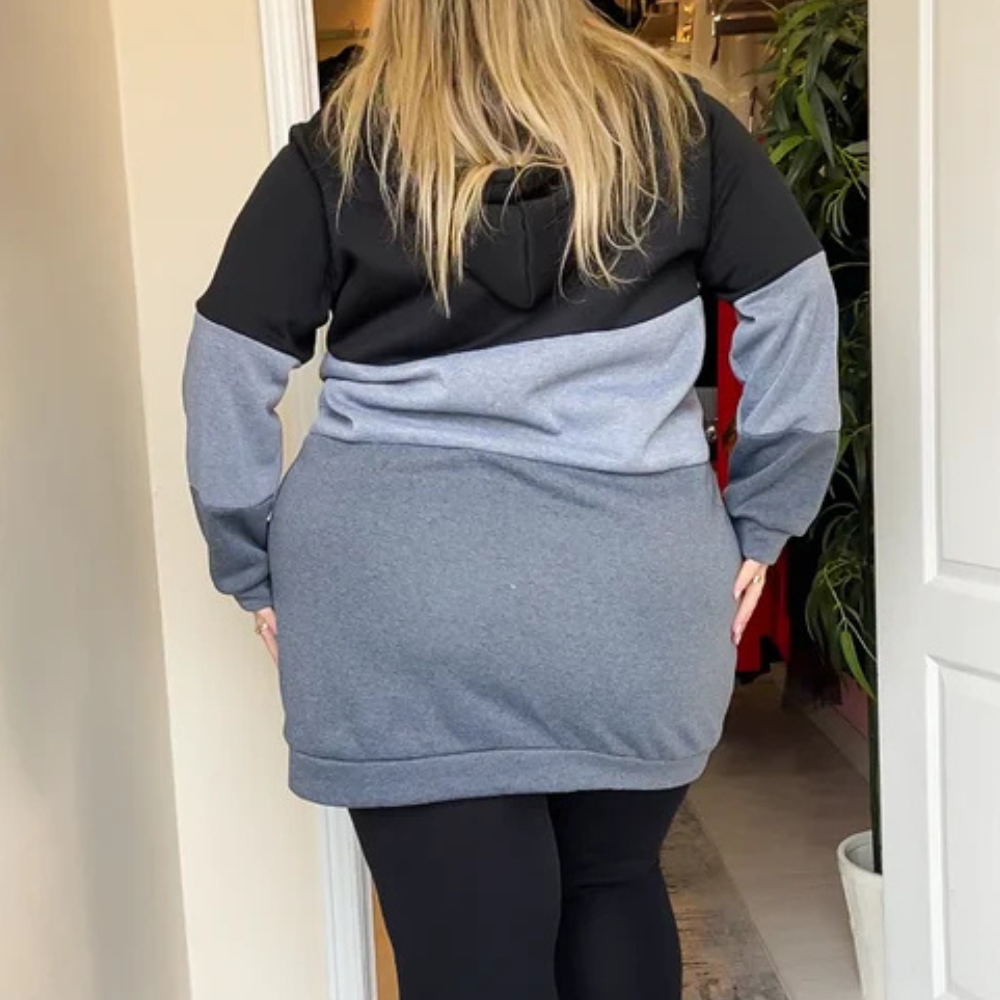 Vrouw in een lange, tweekleurige hoodie met capuchon, zwart en grijs, binnen in een winkel. Comfortabele damesmode, casual kledingstijl.