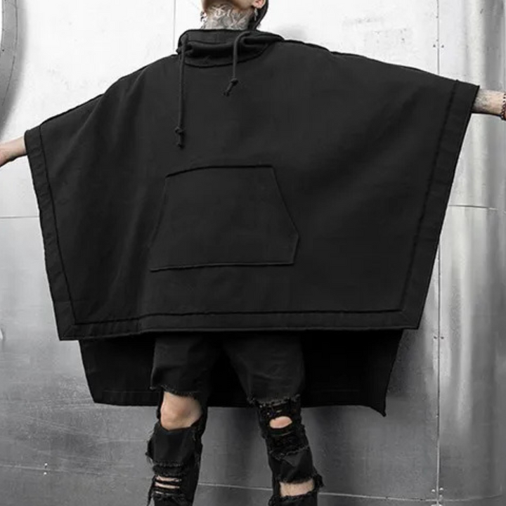 Zwarte oversized poncho met capuchon, grote buidelzak en ruwe afwerking, gedragen door persoon met gescheurde zwarte jeans voor industriële achtergrond.