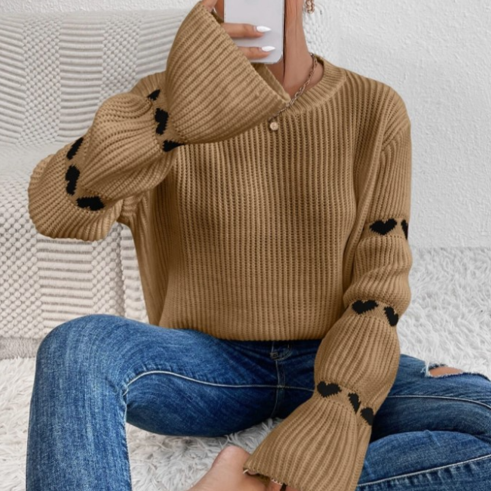 Vrouw in bruine trui met hartjesmotief, zittend op bed met sprei, draagt blauwe jeans. Trendy herfstmode, casual outfit, comfortabele kledingstijl.